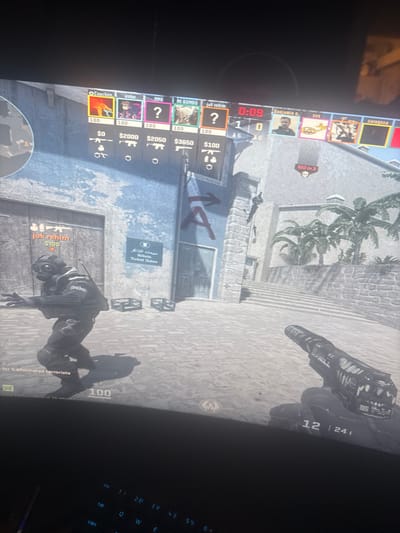 The image displays a computer monitor showing an active first-person shooter video game, observed to be played in the city of Yeroskipou, Cyprus. The in-game scene depicts an outdoor urban environment with light-colored stone buildings and cobblestone paving.
In the foreground, the player's right hand and a black and white weapon, identifiable as a Desert Eagle pistol, are visible. An ammunition counter "12 | 241" is displayed below the weapon. To the lower-left, a third-person perspective shows a character in dark tactical attire, wearing a gas mask and holding a rifle, moving in a crouched or low stance. Above this character, the in-game name "jok-rehim" and a money value "$100" are present.
The background features weathered stone architecture, including a building with a large red "A" and an arrow painted on its facade, indicating a game objective. Below this "A," a blue sign with Arabic text and "Biteb Ticket Sales" in English is affixed to the wall. Two dark-colored crates are positioned on the ground near the base of the building. To the right, a wide set of stone steps ascends, and several green palm trees are visible beyond the structures. A figure is seen clinging to the side of the building on the right.
The top section of the screen displays the game's user interface. A roster of player names includes "Coachen," "oskar," "MMB," "MI BOMBO," "jok-rehim," "Benjamin n.," "SSS," and "Saragosa." Health indicators all show "100." Monetary values for the first five players are "$0," "$2000," "$2050," "$3650," and "$100," respectively. A game timer reads "0:09," and the current score is "1 | 0." Additional interface elements include a prompt "100 in 3" on the right and "for 5 eliminated terrorists" text in the lower-left.