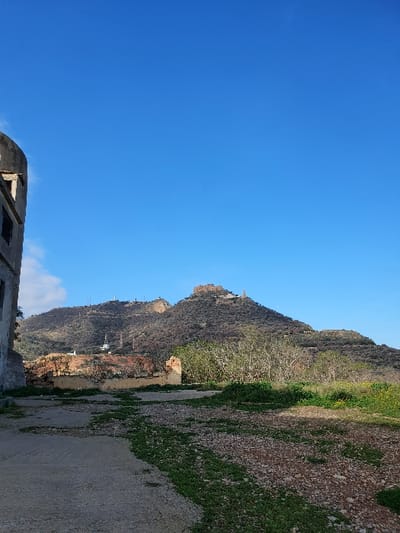 The image shows a hill with a clear blue sky above. The hill is covered in vegetation and appears to be a rocky hillside with a crumbling building on the right side of the frame. It is a clear, sunny day. The location, based on the coordinates provided, is in the city of Oran, Algeria. The image does not include any people or objects other than the hill, the building, and the sky.