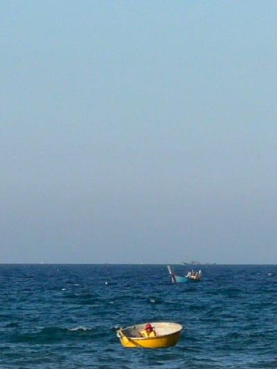 A person wearing a yellow life vest and a red hat sits in a small, yellow, circular boat. The boat is floating on deep blue water. In the background, a larger, blue boat with several people on it is also on the water. Further in the distance, a hazy horizon is visible, with several other boats or ships on the water. The sky is a pale, hazy blue. The scene appears to be the coast of Da Nang, Vietnam, possibly near the Han River or the sea.