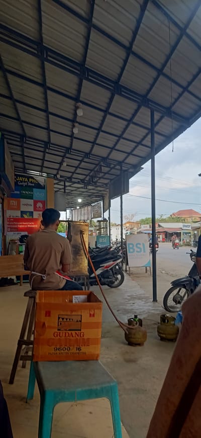 This media file depicts an outdoor scene in Gampongteungoh, Indonesia, under a metal roof supported by dark pillars. The time of day appears to be late afternoon or early evening, given the diffused light and the bright sky visible in the background. A man wearing a light brown shirt and an apron is seated on a blue plastic stool, with his back to the camera. In front of him, on a wooden stool, sits a large cardboard box with "GUDANG GATELUM 9600/16 FILTER KRETEK CIGARETTES" printed on it. A hose extends from the box to a tall, metallic cylindrical object, suggesting some kind of filling or processing activity. Nearby, on the ground, are two gas cylinders.
The setting appears to be a commercial area or a roadside shop, indicated by the storefront visible on the left with signage for "JAYA PHU Gadget Store" and advertisements for various phone brands like Samsung, Oppo, Vivo, Xiaomi, and Advan. On the right side of the street, a sign for "BSI BANK SYARIAH INDONESIA" is prominently displayed. Several motorcycles are parked along the sidewalk, and a few buildings can be seen in the distance, including one with a red roof. A man on a motorcycle is riding past in the background, suggesting some street activity. The overall atmosphere seems casual and functional, with no strong emotional cues conveyed by the subjects. The presence of gas cylinders and a cigarette box hints at a business operation related to these items.