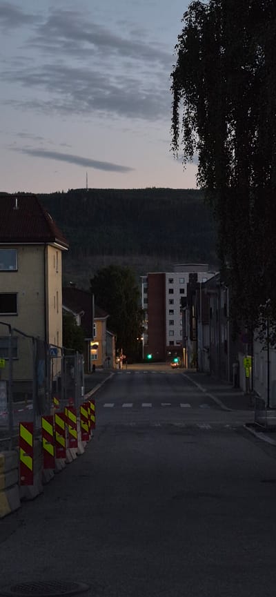 The image depicts a street scene in Lillehammer, Norway, likely during the late evening or early morning given the low light. The perspective is from an elevated viewpoint looking down onto the street and surrounding buildings.

In the foreground, on the right side of the image, several construction barriers with alternating red and yellow reflective stripes are stacked along the edge of the asphalt road. The road itself has white dashed lines marking lanes.

On the left side of the road, a dense line of trees forms a dark backdrop, separating the street from a steep, forested hillside that rises behind it. The sky above is overcast with wispy grey clouds, suggesting a cloudy day or twilight.

Several buildings are visible along the street. Some appear to be multi-story apartment buildings with numerous windows. One building in the mid-ground has a distinctive red tiled roof. Further down the street, illuminated windows suggest some activity or residents within the buildings.

A traffic light is visible hanging over the road, showing a green signal, indicating that traffic is currently permitted to proceed. There are no clear signs of people in the image, and the overall atmosphere is quiet and still, consistent with the time of day. The presence of the construction barriers implies ongoing road work or maintenance in the area. The dense forest on the hillside and the style of architecture are consistent with a Scandinavian setting like Lillehammer.