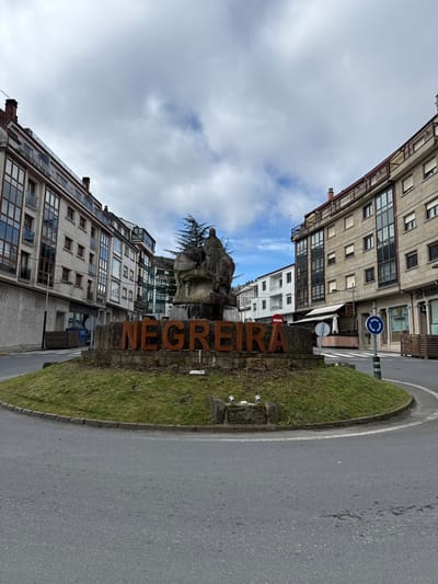 The image depicts a street-level view of a roundabout located in Negreira, Spain, under a partially cloudy sky. The central feature of the roundabout is a circular grassy median containing a large, rusted metal sign spelling "NEGREIRA". Behind this sign, a stone fountain or monument features a sculptural group, including at least one robed figure possibly seated on an animal, with water visibly flowing over the stonework. A small evergreen tree is situated behind the monument.

The roundabout is paved with asphalt and appears clear of traffic. Multi-story buildings flank the roundabout on both the left and right sides. The buildings exhibit varying architectural styles, with multiple windows and balconies; some structures appear residential, while ground floors likely house commercial establishments. On the right side, an outdoor seating area with wooden privacy screens is visible, along with a blue circular road sign indicating a mandatory roundabout direction and a red circular 'no entry' sign further in the background. No individuals or vehicles are prominently visible within the frame.