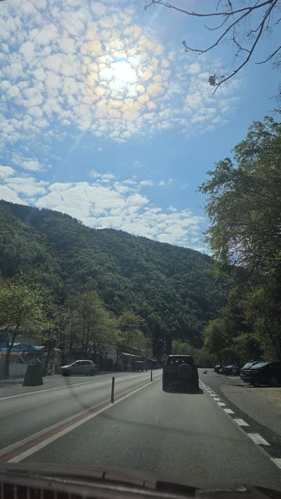 The photograph is taken at street level and is a vertical perspective with landscape features. The sky takes up about half of the frame on the left and is filled with patchy clouds illuminated by the sun. The right side of the frame shows the side of a road and a lush mountainside. Several parked cars are visible on the right side of the road. The photo was taken in Stara Kresna, Bulgaria.