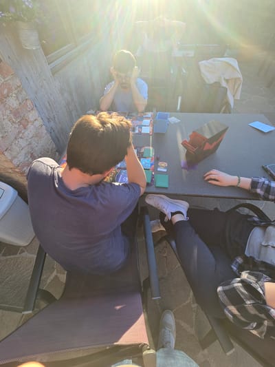 This image was likely taken outdoors in Schwechat, Austria, due to the sun's position and the type of paving stones present. Three individuals are seated around a table. Playing cards are scattered across the surface, with card holders and various gaming accessories visible. One person on the left is resting their head on their hands in a pensive pose. A second person is wearing a grey shirt, and the third person has on a grey cardigan and black and white plaid jacket. The table is set on a stone-paved patio. There is an outdoor chair with brown woven seating visible on the right. The sun is partially obscured, but its glare is still visible in the top-left corner.