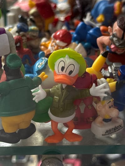 A close-up shot depicts a collection of toy figurines crowded together within a glass-enclosed space. The figurines include a cartoon duck character wearing a green hat and military-style jacket, a Pikachu, and an Asterix figurine, among others. The location is indeterminable.