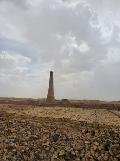 This image, captured in Kuchlak, Pakistan, presents an expansive industrial landscape centered around a brick manufacturing site. A tall, weathered industrial chimney, made of concrete or brick and tapering towards the top, stands as a prominent focal point slightly left of the image's center.

Vast quantities of bricks dominate the foreground and mid-ground. To the right, large, irregular piles of loose, reddish-brown bricks, many appearing broken or older, create a rugged texture. Extending further back and to the left, numerous rows of neatly stacked, yellowish bricks suggest recent production. The ground beneath the bricks appears dry and dusty.

In the distant background, a range of hazy hills or mountains stretches across the horizon. The sky above is largely overcast, filled with various shades of grey and white clouds, though some lighter patches hint at breaks in the cloud cover. The overall lighting suggests it is daytime, under conditions that are either cloudy or hazy. No people are visible, but the sheer scale of the brick piles and the presence of the chimney strongly indicate ongoing or recent industrial activity related to brick making. There is no visible text in the image.