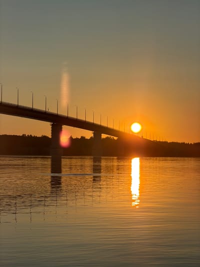 A bridge spans a body of water at sunset. The sky is a gradient of pale blue to warm orange, with the sun a bright, glowing orb positioned above the horizon. The bridge's silhouette is dark against the sky, with a row of streetlights visible along its length. The water reflects the vibrant colors of the sunset, creating a shimmering, golden path. A line of trees forms a dark band along the far bank. The scene is peaceful and serene, evoking the end of a day.