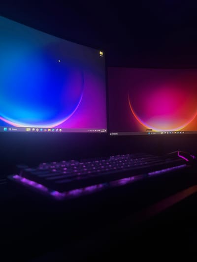 In a dimly lit room, two computer monitors display abstract wallpapers featuring glowing orbs against dark backgrounds. The left monitor shows a vibrant blue and purple orb, while the right monitor features a pink and orange orb. A keyboard with purple backlighting is positioned in front of the monitors, with a purple-lit computer mouse visible on the right side of the keyboard. The overall ambiance is dark and focused on the illuminated technology, suggesting a gaming or work setup. There is no indication of the city of Lormont, France, or any specific location within it.