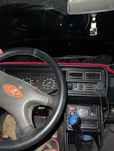 The image shows the interior of a car, likely a Lada, with the steering wheel and dashboard in focus. The dashboard displays a speedometer, tachometer, fuel gauge, and other indicators. A red-trimmed mat is visible on the dashboard. A blue and black gear shift knob stands out in the center console, next to a car stereo. A small air freshener hangs from the rearview mirror. The scene is dimly lit, suggesting it is either nighttime or inside a dark garage. The car is possibly located in Mukur, Kyrgyzstan, based on the user's prompt.
