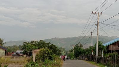 The image depicts a street scene in Blangtingkeum, Indonesia. A paved road stretches into the distance, flanked by vegetation, houses, and utility poles. A motorcycle with a person is traveling along the road. In the background, a hazy mountain range is visible under a cloudy sky. A variety of buildings appear alongside the road, as well as fencing and shrubbery. The overall impression is one of a rural or semi-rural area.