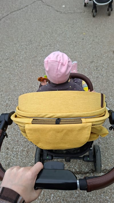 A first-person perspective shot shows a hand gripping a stroller handle as it moves across a paved surface, possibly a sidewalk or park pathway. The stroller is occupied by a young child wearing a pink hat and a dark sweatshirt adorned with small, colorful dots. The child is holding a small toy. The stroller itself features a prominent yellow fabric cover. In the background, another stroller is partially visible. The overall environment suggests an outdoor urban setting. The specific location in Chaikovsky, Russia cannot be determined from the visual information provided.