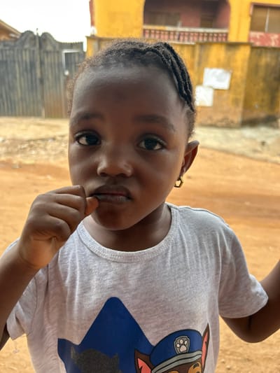A medium close-up photograph captures a young child standing outdoors in Ashipa, Nigeria. The child, of apparent African descent, has dark skin, dark eyes, and dark braided hair. They wear a light grey short-sleeved t-shirt featuring a blue graphic of a character resembling a police dog in uniform. A small, gold-colored hexagonal earring is present in the child's right ear. The child's left hand is raised, with fingers touching the lower lip area. The background consists of reddish-brown dirt ground. To the left, a dark, ornate metal gate is partially visible. To the right, a multi-story building with a yellow-orange facade is present, featuring a reddish-brown balcony structure on the upper level. White rectangular objects are affixed to the wall below the balcony. The scene is illuminated by natural daylight.