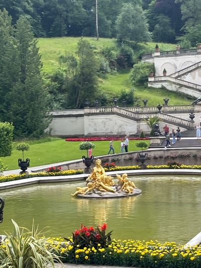 The image depicts a golden fountain sculpture in a large pond, situated within a manicured garden. The sculpture features a central figure with several cherubic figures surrounding it. The pond is bordered by flowerbeds filled with yellow and red blossoms, with lush green grass and trees in the background.
Dominating the upper portion of the image is a grand, ornate staircase leading up a hillside. The staircase is flanked by white balustrades and decorative urns. Several people are visible walking on the steps and on a pathway at the base of the staircase.
The overall setting suggests a formal garden or park, likely associated with a historical or royal residence. The vegetation is verdant, indicating a temperate climate during the time the photograph was taken.
The location is identified as Ettal, Germany. This likely corresponds to the gardens of Ettal Abbey, a Benedictine monastery. The architectural style of the staircase and the fountain's classical theme are consistent with European Baroque or Rococo garden design, often found in such historical sites. The image captures a serene and picturesque scene, showcasing the interplay of sculpture, water, flora, and architecture.