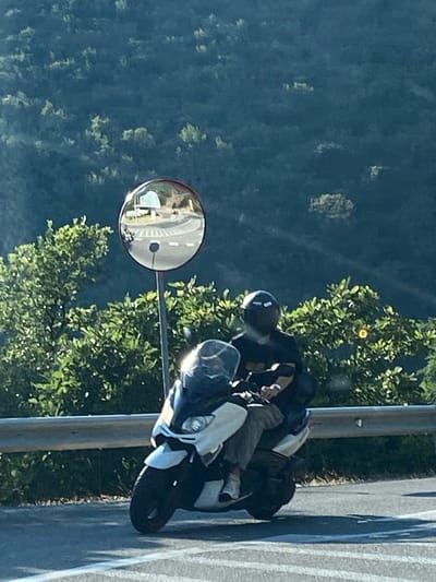 Two individuals, both wearing helmets, are riding a white scooter on a paved road. The driver is wearing a black shirt and grey pants, while the passenger is wearing a black shirt and beige pants. The scooter has a windshield and is positioned on the right side of the road, with a guardrail to its left. In the background, lush green trees cover a hillside, and a convex mirror is mounted on a pole, reflecting the road and vehicles. The scene appears to be located in Novoselje, Montenegro.