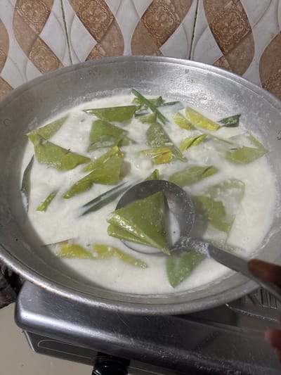 In a domestic kitchen in Blangseureukuy, Indonesia, a metal wok filled with a creamy white liquid and green leafy vegetables is being stirred with a metal ladle. The wok sits on a gas stove. The background features a wall tiled with a diamond pattern in shades of beige and brown.
