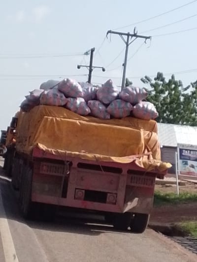 A red flatbed truck is overloaded with sacks on top of a yellow tarp. The sacks are patterned in red and white. The truck is driving on a paved road. Overhead power lines and poles are visible above the truck. To the right of the truck is a tree and a sign with the text "T&T MED". The image appears to be taken in Dungu, Ghana.