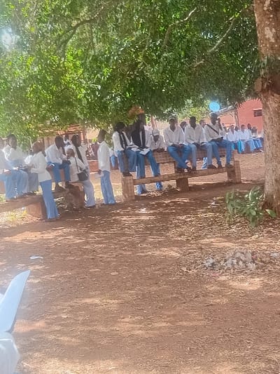 A line of seated individuals in white shirts and blue pants or skirts, possibly students, are visible in the image. They are sitting on a wooden structure in an outdoor setting. The area is under a tree, and the ground appears to be dirt or sand with some debris scattered around. A building is visible in the background. The photo is taken from a slightly elevated angle and the orientation is rotated.