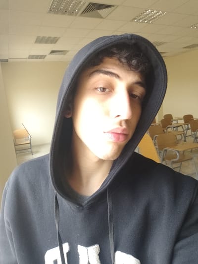A close-up, chest-level selfie depicts a young male subject in what appears to be a classroom or lecture hall in Jibb as-Safa, Syria. The individual, who has dark, curly hair and brown eyes, is wearing a black hooded sweatshirt with an unidentifiable white graphic design visible on the chest. The hood is pulled over his head. His face is partially illuminated from the left, creating a shadow on the right side. His gaze is directed forward, towards the camera.

In the background, on the right, several rows of empty student desks with light-colored wooden tops and metal frames are visible. To the left, a plain, light-colored wall is present, with a single wooden chair with a metal frame positioned against it. The ceiling features a grid of white acoustic tiles, inset fluorescent light panels, and a ventilation grille. The room appears brightly lit, with strong illumination originating from the left side of the frame.