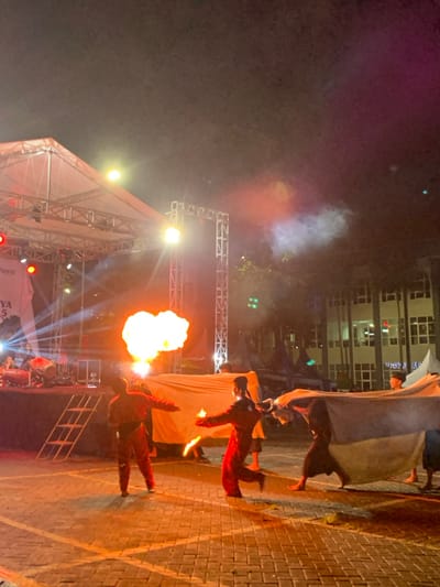 The scene takes place at night in front of a stage in the city of Kecamatan Batu, Indonesia, at the coordinates -7.867 latitude and 112.514 longitude. Five men wearing dark, red, or blue clothing are performing a fire dance in front of a large white sheet. There are red and blue lights illuminating the scene, and a fire is burning, creating a large orange flame. The men are moving their bodies in a coordinated fashion as they maneuver around the fire. The ground is made of brick pavers. There is a ladder on the left side of the scene, which is used for access to the stage, where a large white sheet is suspended. There are buildings in the background, which are lit up. The image appears to have been taken at a festival or other event.
