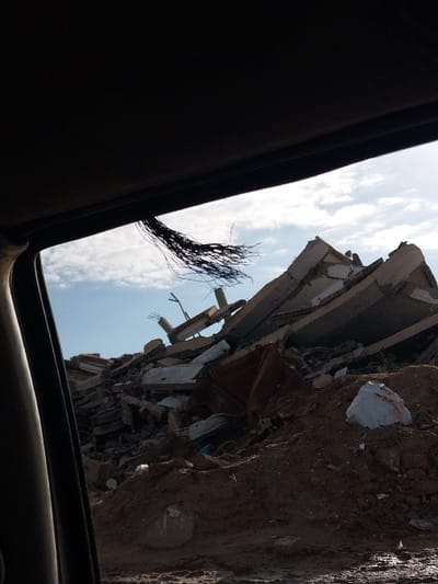 The image, taken from within a vehicle, depicts a large pile of building demolition debris in a-Zawayda, Palestinian Territory. The upper section of the frame shows the dark interior roof lining and window frame of the vehicle, from which a cluster of dark, thin, fragmented strands, resembling wires or rebar, hangs from the upper left. Beyond the vehicle's interior, a substantial mound of rubble occupies the midground, consisting of fractured concrete slabs, exposed reinforcing steel bars, and various shattered building materials. This debris pile rests on uneven, reddish-brown earth, which appears damp in certain areas. Directly in the foreground, on the ground, a white, irregularly shaped object is visible on the right, and a partial circular concrete structure is embedded in the soil on the left. The background displays a clear blue sky with scattered white cumulus clouds. No individuals or ongoing activities are visible within the scene.