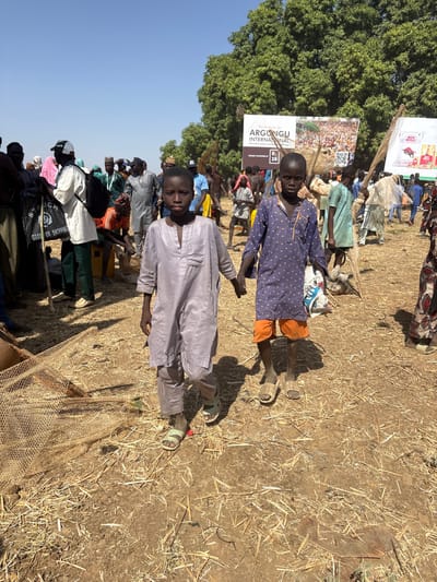 The image depicts an outdoor gathering in Kokani North, Nigeria, under a clear blue sky. In the foreground, two dark-skinned boys, appearing to be adolescents, walk towards the viewer holding hands. The boy on the left wears a light grey long-sleeved tunic and matching trousers, with green sandals. The boy on the right wears a dark blue long-sleeved shirt with light polka dots, orange shorts, and light brown sandals; he carries a white and blue plastic bag in his left hand.

The ground is dry, dusty earth, scattered with straw and sparse dry vegetation. A woven net structure is visible on the ground in the lower-left corner. In the mid-ground and background, a large crowd of individuals, predominantly adults, are visible, dressed in a variety of traditional and casual attire, congregating and moving.

A large white banner in the mid-background reads "Welcome to ARGUNGU INTERNATIONAL" and "R. 28," featuring an image of a crowd and a QR code. To its right, a partially visible white banner displays the "BIG BULL" logo and text. A line of dense green trees forms the far background.