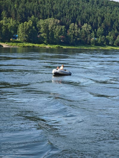 In Pristan', Russia, on a wide, blue river with visible ripples, two people are in an inflatable raft. The raft is gray and has two individuals seated inside. One person is wearing a light-colored shirt and a hat. The other person is also wearing a light-colored shirt. The raft appears to be moving, as indicated by the wake behind it. The background features a lush, green forest that slopes down towards the river, with some buildings and trees visible on the shore. The sky is clear and bright.