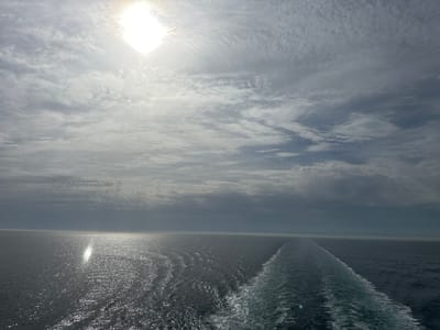 The image depicts a seascape under a partly cloudy sky with the sun shining through the clouds. The body of water, likely an ocean or sea, is relatively calm with gentle waves. A trail of white foam suggests a vessel has recently passed, creating a distinct wake. The sun reflects on the water's surface, forming a bright specular highlight. The location is not discernible from the image.