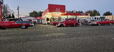 The image depicts an outdoor gathering at dusk in Centralia, Washington, specifically at 122 North Tower Avenue, in front of a commercial building identified as RI WRIGHT ROASTERS. The building features a red upper facade and a white lower facade, illuminated by decorative string lights.
On the asphalt pavement in the foreground and mid-ground, a display of classic American automobiles is present. From left to right:
1. A red and grey two-tone 1955-1957 Chevrolet Bel Air two-door post sedan, equipped with aftermarket chrome wheels, is parked facing right.
2. Centrally, in front of the building, a dark red custom 1930s Ford coupe hot rod with exposed engine components (multiple carburetors/scoops) and black wheels is parked.
3. To the right, a red 1960s-era four-door sedan, consistent with a Chevrolet Impala, also with chrome wheels, is parked.
4. Another darker classic car is partially visible behind the red sedan.
Approximately fifteen individuals are dispersed throughout the scene, primarily observing the vehicles or engaging in conversation. Near the main building's entrance, a microphone stand and a large speaker are set up, indicating a live performance or announcement. A red pop-up canopy tent is erected on the left side of the building. In the far right background, a large white recreational vehicle (RV) is parked. The sky displays a pale blue-grey hue, characteristic of evening, with power lines and utility poles visible against it.