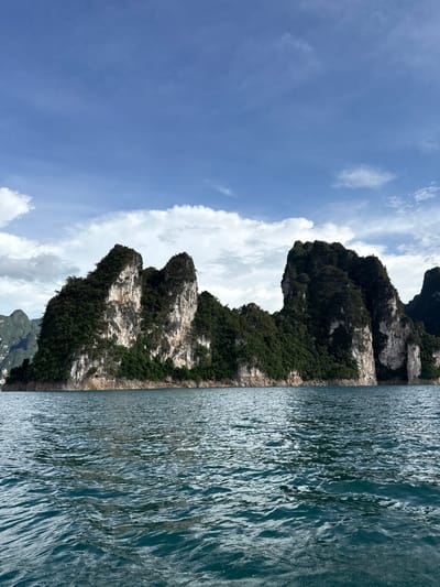 A wide, horizontal shot captures a scenic view of limestone karst islands emerging from a body of water under a partly cloudy sky. The islands are densely covered in lush green vegetation, with sheer, rocky cliffs showing in patches. The water in the foreground is a deep blue-green, showing gentle ripples and reflections. The sky above is a bright blue, dotted with white, fluffy clouds. The scene is reminiscent of Cheow Lan Lake in Khao Phang, Thailand, known for its dramatic karst formations. No people or specific actions are visible, creating a serene and natural landscape.