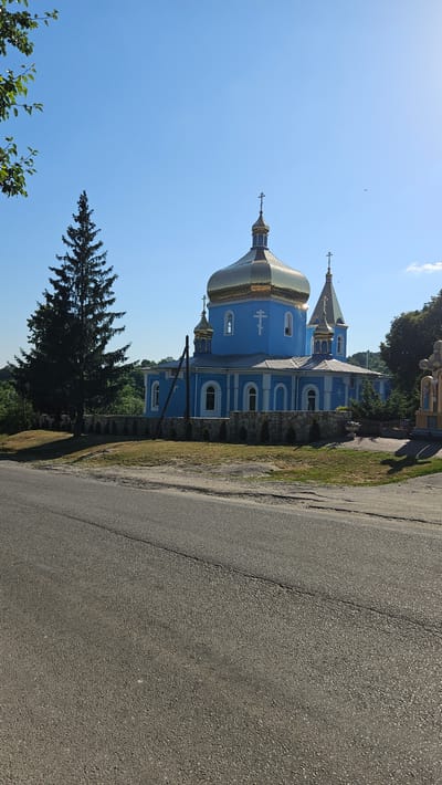 The image depicts a blue Eastern Orthodox church with golden domes. The church has a prominent central dome and a smaller dome on the left, both topped with crosses. The building features arched windows and decorative elements. It is situated in an outdoor setting, with a paved road in the foreground and a grassy area with some trees and foliage to the side and behind the church. A tall evergreen tree stands in front of the church. The sky is clear and blue, suggesting a sunny day. The orientation of the image is rotated 90 degrees clockwise. There is no visible text in the image. The scene appears to be in a rural or semi-rural area. The time of day is likely daytime, given the bright lighting and clear sky.