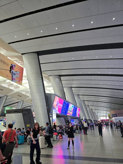 This media file captures a bustling scene within a modern, spacious interior, likely a train station or airport in Fengtai District, China. The architecture is striking, with a high, vaulted ceiling composed of textured panels and integrated lighting. Large columns support the structure, and the walls feature a repeating pattern of paneling.
The primary subjects are numerous people, many of whom are seated or standing in queues, suggesting they are waiting for transportation. Luggage, including suitcases and bags, is visible throughout the scene, further indicating travel. A few individuals in dark uniforms, possibly security or station staff, are also present.
A prominent feature is a large digital display screen showcasing vibrant graphics and text, contributing to the modern aesthetic. Another large banner or advertisement is visible on a lower level, depicting a person in athletic wear.
The lighting suggests it is daytime, with artificial lights from the ceiling and the digital displays creating bright, colorful accents on the floor. The atmosphere appears orderly but busy, with individuals engaged in waiting and moving about. The overall impression is one of a well-trafficked public transportation hub. Some visible Chinese text is present on signage and advertisements, contributing to the location context.