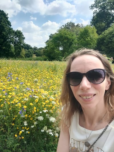 The image shows a woman taking a selfie in a field of yellow and blue wildflowers. She is wearing sunglasses, a white t-shirt, and a silver necklace. The background is a blur of green trees and a blue sky with white clouds. The location is a meadow in Isselburg, Germany.