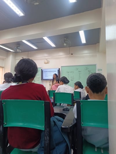 The image depicts an indoor classroom setting, likely in Quezon City, Philippines, characterized by fluorescent lighting strips on the ceiling and ceiling fans. In the foreground, the backs of students are visible, seated in green plastic chairs. The students are facing towards a presenter, a woman in a light brown top, who is standing in front of a whiteboard and a projector screen. The projector screen displays text about "Exceptions to Informed Consent" with bullet points, while the whiteboard has handwritten notes and numbers. The overall atmosphere suggests an educational lecture or presentation in progress.