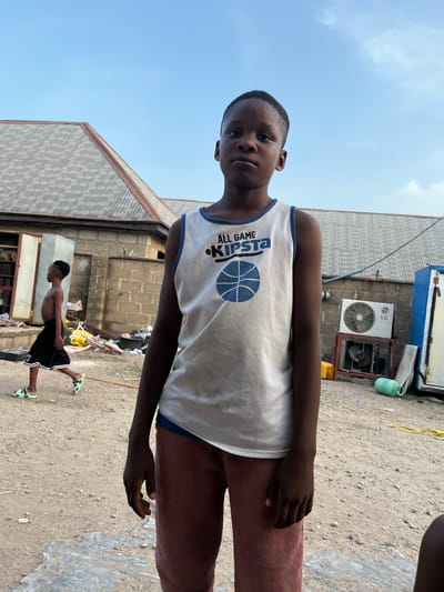 A young boy in a white tank top with the word "KIPSTA" and a basketball graphic stands facing the camera. He wears reddish-brown corduroy pants. Behind him, a second shirtless boy in shorts and sandals walks away. The background consists of buildings, including one with an LG air conditioner, and a dirt ground with debris. The scene appears to be located outdoors. The location of the image is Iddah, Nigeria.