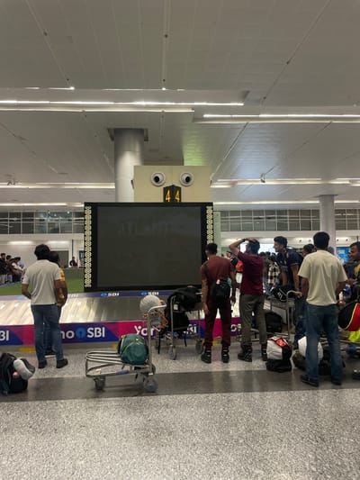 The image depicts a busy airport baggage claim area. In the foreground, several individuals are standing near a baggage carousel, waiting for their luggage. Luggage carts are visible, some with bags placed on them. A large digital screen displays information, with the number "44" prominently shown. The environment appears to be an indoor terminal with a high ceiling, fluorescent lighting, and tiled flooring. The overall scene suggests a common travel scenario at an airport. The location is identified as Raipur Khurd, India.