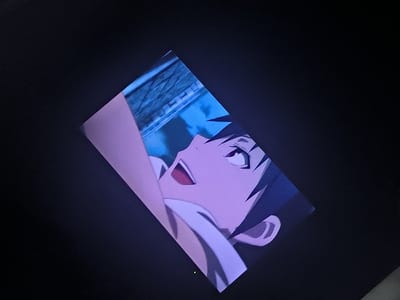 The image shows an anime character with dark hair and bright eyes, appearing to look upwards with an open mouth, possibly expressing surprise or excitement. Part of his ear and neck are visible. The background has a blurred texture that resembles a fence or some architectural element under a bright blue sky, suggesting an outdoor scene. The image is presented at an angle on what seems to be a digital screen or monitor.