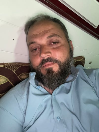 The image shows a man lying on his side, possibly on a bed or sofa. He has a beard with some gray hairs and appears to be looking directly at the camera. The man is wearing a light blue shirt with buttons. The background is blurred, but it appears to be an indoor setting. There are some abstract patterns visible in the lower right portion of the image, possibly from a pillow or blanket. The lighting suggests it could be daytime, but there are no specific cues about the time of day or weather conditions. The man's expression is neutral. There is no visible text in the image. Given the location context of Kabul, Afghanistan, the scene is likely domestic. The man's pose and expression could suggest relaxation or rest.