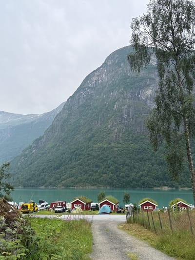 The image shows a camping area near a lake in Olden, Norway. In the foreground, a gravel path leads towards a row of small, red cabins with green, grass-covered roofs. Several tents are pitched near the cabins, and a fence made of wooden posts and wire runs along the right side of the path. Beyond the cabins and tents, a bright turquoise lake is visible, with a large, green, tree-covered mountain rising steeply from the opposite shore. The sky is overcast and a tall tree with delicate leaves stands on the right side of the image, partially obscuring the mountain. In the middle ground, several vehicles are parked, including a yellow camper van, a white camper van, and a silver car. A few people are seen near the vehicles and cabins. The overall scene depicts a serene, natural setting with a focus on outdoor recreation.