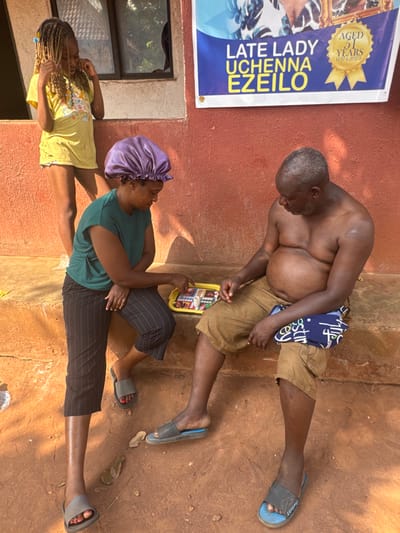 The image depicts an outdoor scene in Aguata, Nigeria, in front of a reddish-brown plastered building. Three individuals are present. On the left, a young female with blonde braided hair, wearing a yellow t-shirt and shorts, stands observing. In the center, an adult female with a purple satin bonnet, dark green t-shirt, striped capri pants, and grey slide sandals is seated on a low concrete step. Her right hand is positioned over a Ludo board game set on a yellow tray, indicating engagement with the game. To her right, an adult male, bare-chested with a prominent abdomen, wearing light brown cargo shorts and dark slide sandals, is also seated on the step, looking down at the game. A blue and white patterned cloth is draped over his left knee. Above the scene, a blue and white banner is affixed to the wall, bearing the text "LATE LADY UCHENNA EZEILO" and "AGED 51 YEARS (1974-2025)". The immediate foreground consists of reddish-brown earth with scattered leaves and small stones.