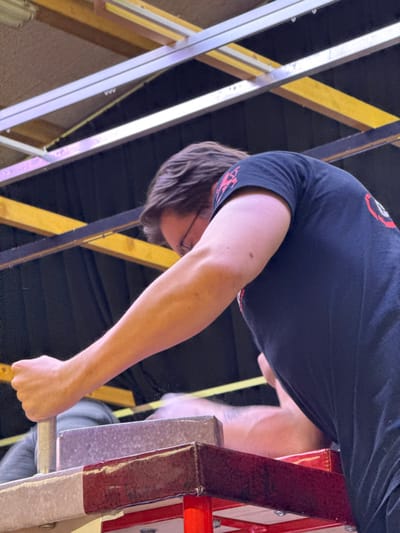 An indoor arm-wrestling competition is depicted in Reisenberg, Austria. The image captures a male participant, viewed from a low angle, leaning forward over an arm-wrestling table. He has medium-length brown hair, wears glasses, and is dressed in a dark short-sleeved t-shirt featuring a red and white logo on the right shoulder area. His right arm is extended, and his right hand firmly grips a vertically positioned, ribbed metallic handle on the arm-wrestling table.
The arm-wrestling table is prominent in the foreground, characterized by padded edges in red and white sections and a grey-toned upper surface. To the right of the metallic handle, a blurred human forearm and hand, belonging to an opponent, are partially visible on the table, indicating an ongoing match. A third individual, wearing a grey hooded garment, is vaguely visible in the blurred background to the left, behind the table. The overhead environment features a ceiling with visible yellowish-brown wooden beams and silver metallic support structures set against a dark background, suggesting a functional indoor venue with artificial illumination.
