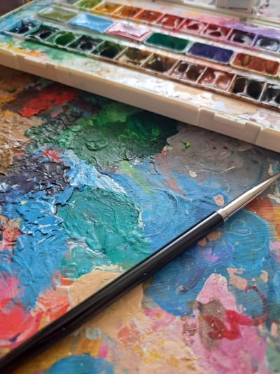 This is a close-up, overhead shot of an artist's palette covered in thick, textured paint. A black-handled paintbrush with a metallic tip rests diagonally across the palette, its bristles touching the vibrant hues. The palette itself is a rough, wooden surface, worn and splattered with a chaotic yet beautiful arrangement of colors. Dominant colors include various shades of blue, green, pink, red, and brown, applied with visible brushstrokes and impasto techniques.

To the left of the painted palette, a white plastic watercolor paint set is partially visible. The paint wells are filled with an array of colors, some appearing wet and fresh, others dried and well-used. The arrangement suggests a working artist's setup.

The scene is brightly lit, likely indoors in an art studio or workspace, given the close-up perspective and the nature of the subjects. There are no visible signs of the time of day or weather. The overall impression is one of artistic creation and activity. The textures and colors evoke a sense of creativity and passion. There is no visible text in the image. The location context of Popovo, Bulgaria, is provided, but there are no specific visual cues within the image to confirm or suggest this location.