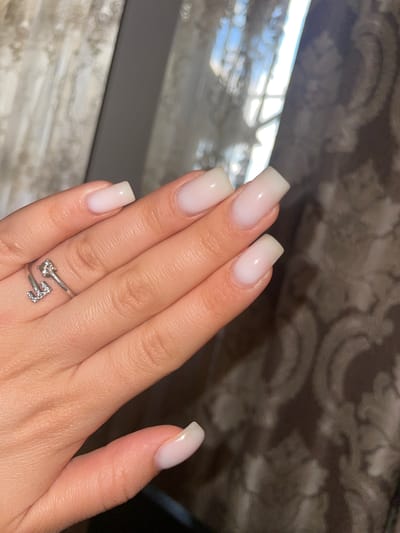 The media file shows a close-up view of a person's hand, adorned with square-shaped, light pink or nude-colored acrylic nails. The hand is positioned on a textured background that appears to be fabric with a patterned design. A silver ring with small, sparkling stones is worn on the ring finger. The ring has a stylized "F" shape made of stones and what appears to be a single stone at the other end of the ring. The lighting suggests an indoor setting, possibly with natural light filtering in from a window, creating reflections on a surface in the background. The overall impression is one of personal grooming and style. No specific activity or event is depicted, but the focus on the nails and ring suggests an emphasis on personal adornment. The location context provided is Armavir, Armenia, but there are no overt visual cues within the image to confirm this specific location. The time of day and weather are not discernible from the image. There is no visible text in the media file.