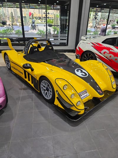 This is a medium-angle, eye-level shot of a bright yellow, low-profile sports car parked indoors on a gray tiled floor. The car is the primary subject and dominates the foreground and midground. It features a carbon fiber hood, black seats with "SR" branding, and a large white "3" on the front. The car has a license plate with Thai characters and the number "U3333." To the left of the yellow car, a portion of a pink car is visible. In the background, through a large glass window, a botanical garden with various cacti and lush greenery can be seen. The text "PATTAYA" is visible on the window. The overall environment appears to be a showroom or exhibition space, possibly at the Nong Nooch Tropical Botanical Garden in Na Chom Thian, Thailand, given the context of the cacti display. There are no people visible in the frame.