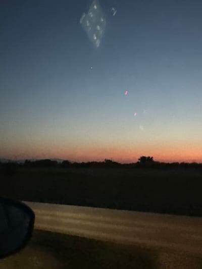 The image depicts a twilight sky over a rural landscape. The sky transitions from a deep blue at the top to lighter shades of orange and pink near the horizon. Silhouetted against the sky are trees and a low-lying landform. In the foreground, a dark, amorphous shape is visible on the left, possibly a car mirror or window. A road or path runs horizontally across the bottom half of the image, with faint stripes of light on its surface. There are a few small, bright specks in the sky, one appearing as a white dot and two others as vertical red lines. A blurry, diamond-shaped reflection is also present in the upper part of the sky. The scene appears to be taken from a moving vehicle. The location of Skutare, Bulgaria is not discernible from the visual information in the image.