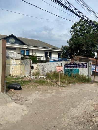 The scene is a dirt road in front of a dilapidated concrete building in the city of Kecamatan Dayeuhkolot, Indonesia (latitude: -6.963, longitude: 107.614). The building appears to be under construction or in a state of disrepair. There is a sign in Indonesian that reads "DIJUAL TANAH SHM" ("For sale land SHM") on a wooden post. There is graffiti on the wall of the building, and several posters are displayed with the text "ON ON ON ON ON ON". A large, black plastic trash bag is lying on the ground next to the building. The scene is largely empty, with no people visible. The sky is overcast with clouds, and there are power lines running across the top of the image. A portion of a green leafy tree is visible on the right side of the image. The dirt road is dusty and unpaved.