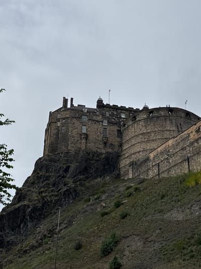 A low-angle, full shot captures Edinburgh Castle, perched atop volcanic rock under a cloudy sky. The castle's stone structure, made of dark, weathered blocks, dominates the upper portion of the frame. Multiple turrets, battlements, and numerous windows are visible. Two flags fly from the castle, one with a red and white design, and another with blue and white. In the foreground, a steep, rocky hillside covered in sparse green vegetation slopes downwards, with a single lamppost visible. The overall atmosphere is somber and imposing, typical of the historic fortress in the heart of Edinburgh, Scotland, United Kingdom.