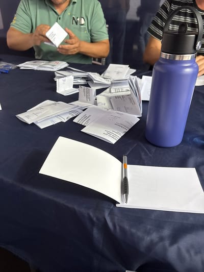 An over-the-shoulder, medium shot captures a table draped with a navy blue cloth, cluttered with numerous white envelopes and papers. In the background, a person wearing a green polo shirt with "ND" visible on the chest is handling a stack of white papers and envelopes. To the right, a purple water bottle with a black lid stands prominently. In the foreground, an open notepad with a pen resting on it suggests an ongoing activity. The scene appears to be an indoor setting, possibly a polling station or an administrative office. Given the mention of "ELECCIONES GENERALES 2026" and "ACCION PARA CEPRESIDENTES" on some of the documents, this scene is likely related to the upcoming presidential elections in Peru, which would place it in a location within the city of Santiago de Surco, Peru, given the provided context.