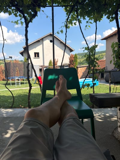 The image depicts a backyard scene in Kirchevo, Bulgaria, on a sunny day. In the foreground, a person's legs and feet are visible, clad in grey shorts, resting on a green plastic chair. The person's feet are crossed. The background reveals a spacious yard with a trampoline and a small blue inflatable pool. A red slide is also visible in the yard. Two houses are partially visible, one a light-colored two-story building with a dark-framed window, and another to the right with a red-tiled roof. Green vines with leaves hang from an overhead structure, casting dappled shadows on the ground and the person's legs. The overall impression is one of leisure and a relaxed atmosphere in a residential setting.