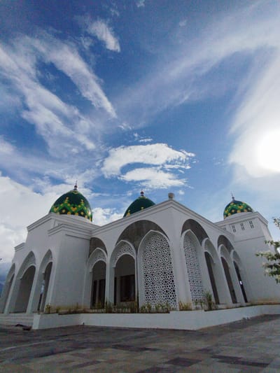 A low-angle exterior shot captures the side of a mosque in Keureuweungkrueng, Indonesia, on a bright, partly cloudy day. The mosque features a white, modern architectural style with a series of arched doorways and windows. Some of these windows have intricate, geometric lattice designs. Three prominent domes top the mosque, two of which are clearly visible. These domes are a vibrant green and yellow checkered pattern. The ground in front of the mosque is paved with dark asphalt. The sky above is a clear blue with scattered white, wispy clouds and a bright sun partially obscured. There are no people visible in the image.