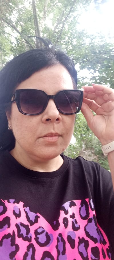 A woman with dark hair and sunglasses is taking a selfie outdoors. She is wearing a black t-shirt with a pink and purple leopard print design. Her right hand is touching the side of her sunglasses. The background is blurred, showing green trees and foliage, suggesting an outdoor, possibly park or natural setting. The lighting is bright, with some overexposure in the upper right corner. The image appears to be taken in the city of Zhirnovsk, Russia.